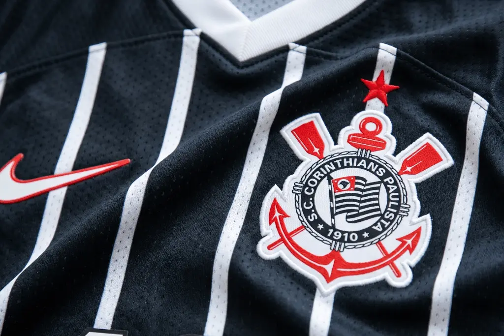 Close-up detalhado da camisa oficial do Sport Club Corinthians Paulista, destacando o escudo bordado com âncora e remos sobre o tecido preto com listras verticais brancas. A imagem representa a identidade visual usada nos confrontos contra o maior rival do Corinthians.