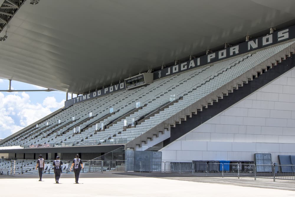 Vista em perspectiva da arquibancada da Neo Química Arena com a frase "Jogai Por Nós" estampada na cobertura. Três funcionários caminham pela área de circulação em frente aos assentos brancos e cinzas.