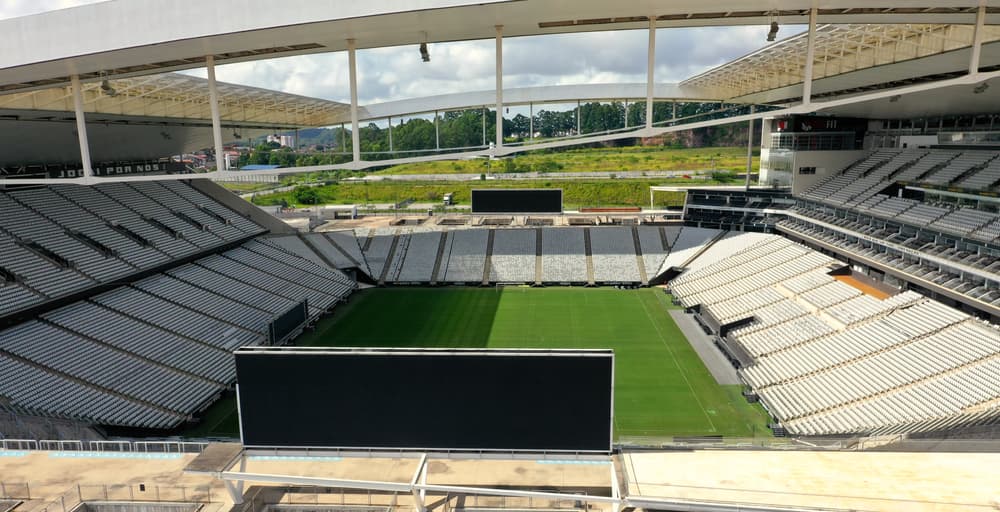 Ampla visão interna da Neo Química Arena destacando a arquitetura moderna da cobertura e o contraste entre as cadeiras brancas e o campo de futebol. Ao fundo, é possível ver a vegetação e o entorno do estádio em Itaquera.