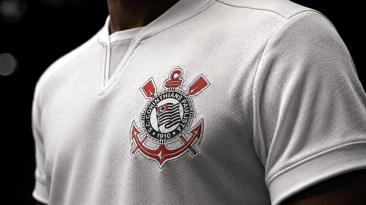 Close no uniforme branco do Corinthians, com escudo em destaque no peito e fundo escuro desfocado.
