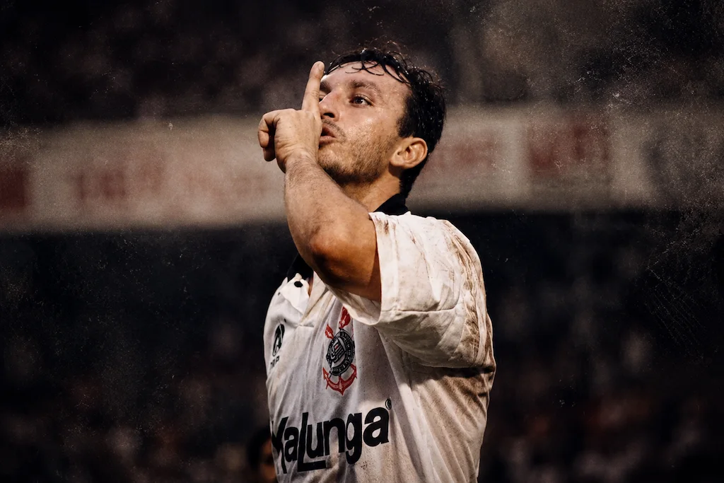 Craque Neto comemora com gesto de silêncio usando a camisa do Corinthians em imagem editorial dramática com atmosfera de estádio e iluminação cinematográfica.